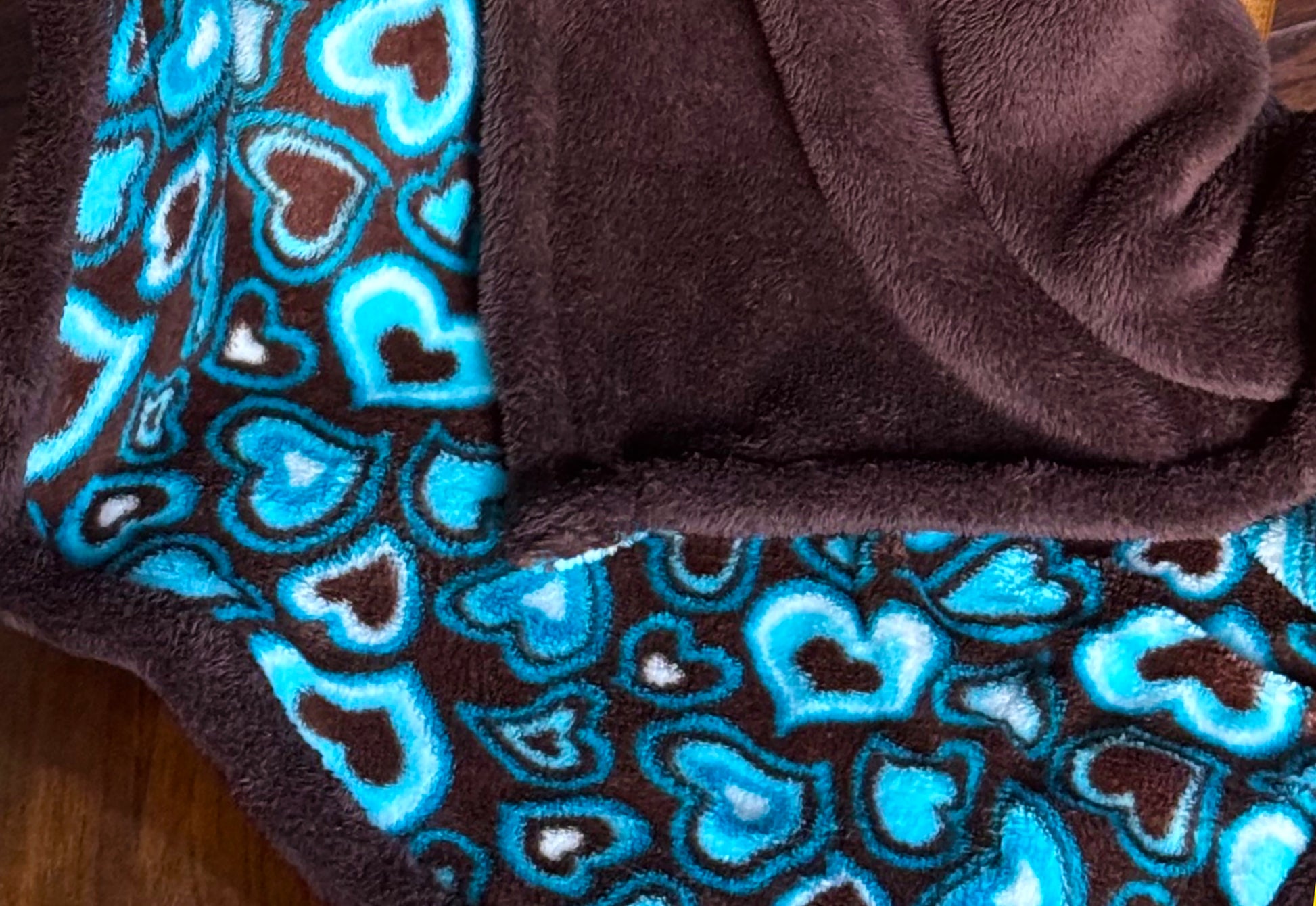 The cuddliest blanket with a pattern of hearts in different sizes and colors of light & medium blue, brown, and white on one side, and solid brown on the opposite side.  Made with up-cycled polyester fabric.  Perfect for those chilly nights with a hot cup of cocoa.  Dimensions: 46" x 36" Machine wash cold, air dry. Made with Love in the USA